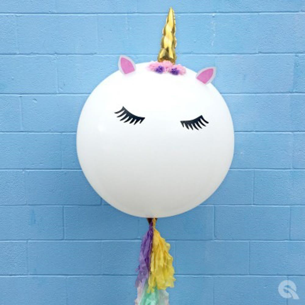 Round white balloon with closed black eyes, pink ears, purple and yellow flowers, and a gold unicorn horn topper.