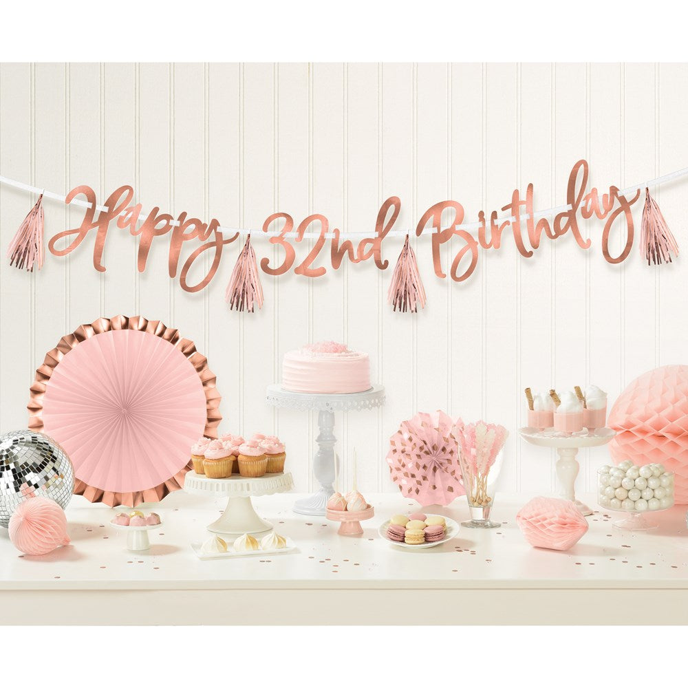 Rose gold foil "Happy 32nd Birthday" banner with pink tassels, paired with pink and white party decor on a white table.