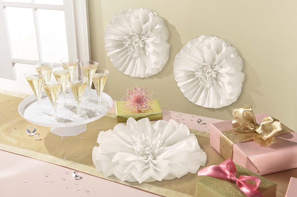 Three white fluffy fabric flowers with layered petals displayed on a table with champagne glasses and wrapped gifts.