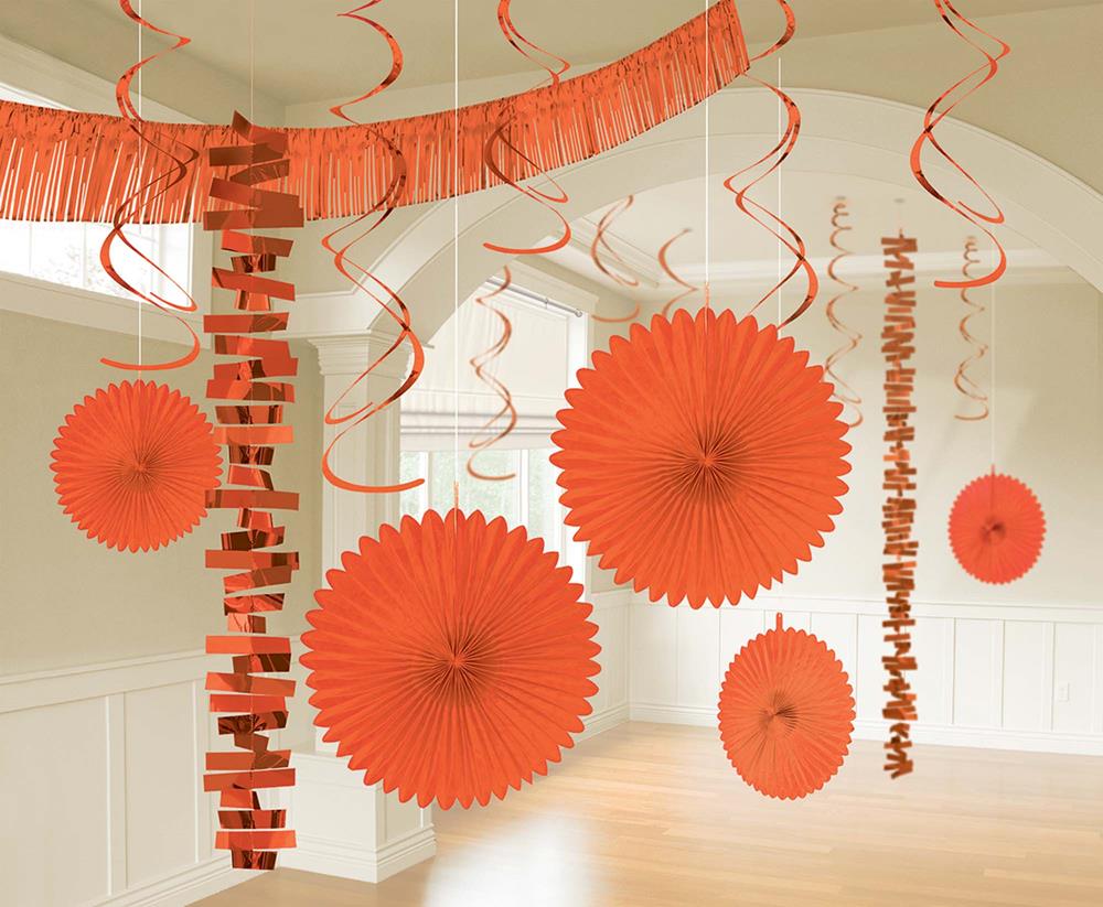 Bright orange hanging paper fans, spirals, and fringe garlands decorating a neutral room ceiling for a festive party.