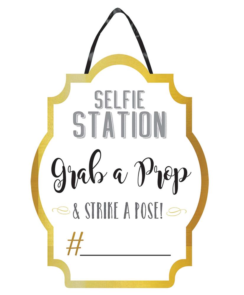 White sign with gold border reading "Selfie Station, Grab a Prop & Strike a Pose!" with a hashtag line at the bottom and a.