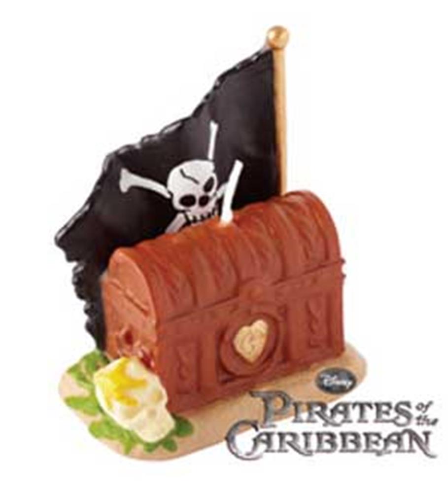 Brown treasure chest candle with pirate skull flag on wooden pole, placed on sandy base with white flower.