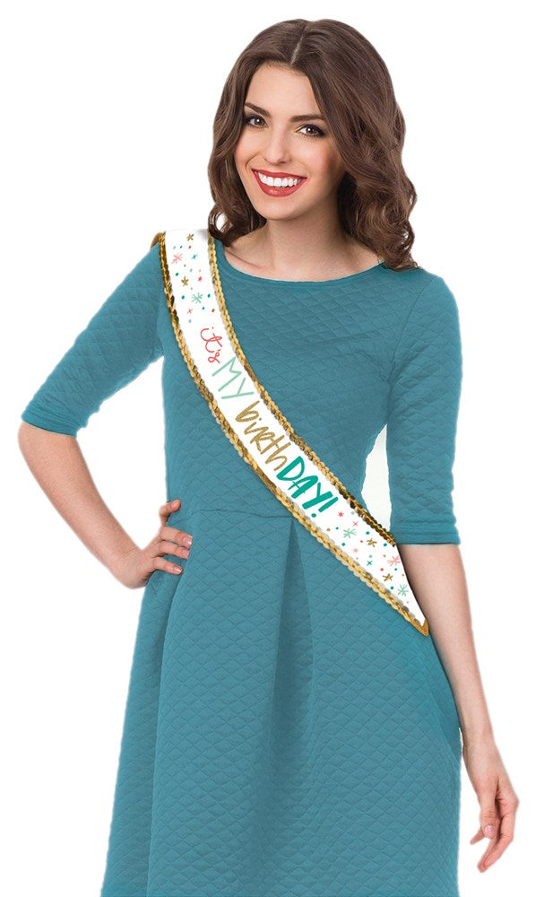 White fabric birthday sash with colorful confetti and gold "Happy Birthday!" text worn over teal dress.