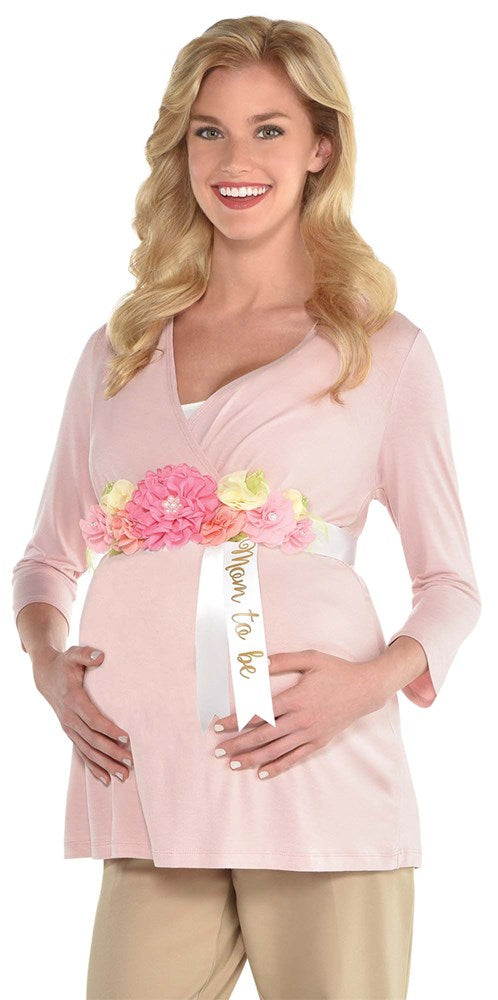 Pink satin sash with floral embellishments and "Mom To Be" gold lettering worn over light pink maternity top.