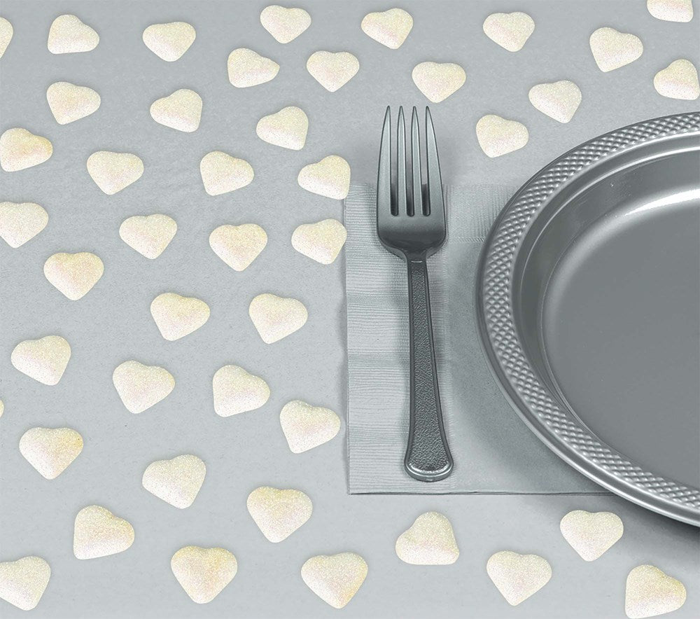 Small iridescent white heart confetti scattered on gray surface next to silver fork and textured silver plate.