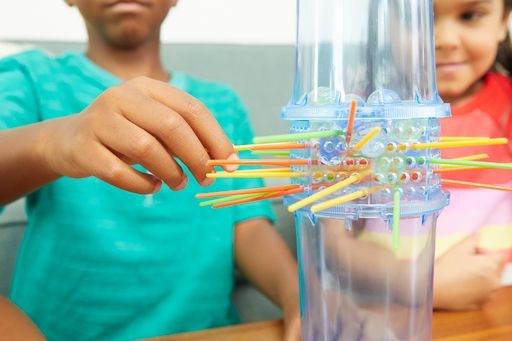 Clear plastic Kerplunk game tower with yellow, orange, and blue sticks inserted horizontally, children playing in background.