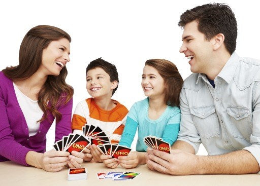 Family of four smiling and holding colorful Uno cards while playing the Mattel Uno Card Game House Rules on a table.