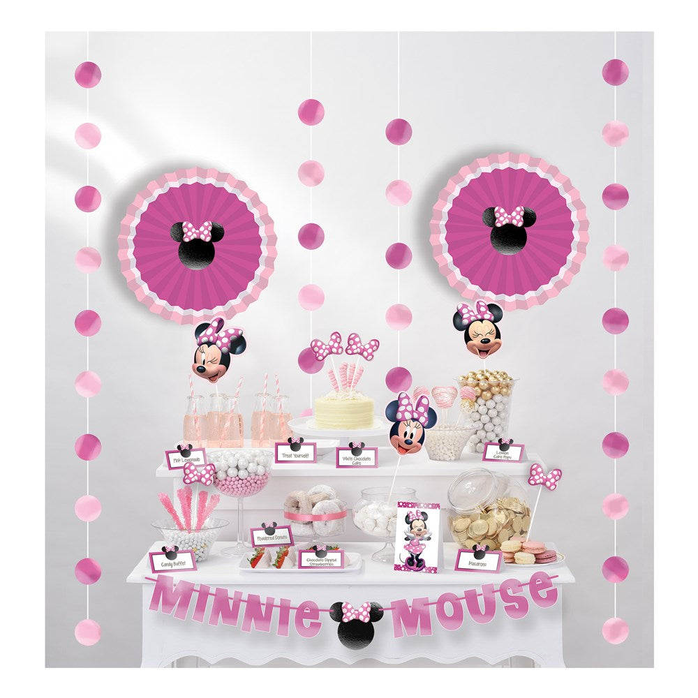 Pink and white Minnie Mouse themed buffet table kit with hanging rosettes, paper cups, popcorn boxes, and a "Minnie Mouse".