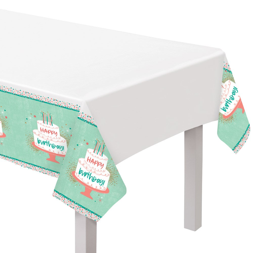Plastic table cover with turquoise border featuring repeating white birthday cakes and "Happy Birthday" text, white center,.