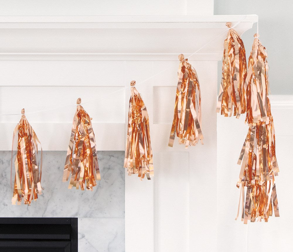 Rose gold foil tassel garland with six metallic tassels hanging on white mantel.