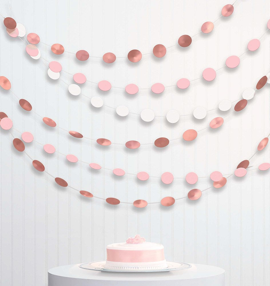 Six strands of circular rose gold, blush pink, and white paper garlands hanging against a white wall with a pink frosted.