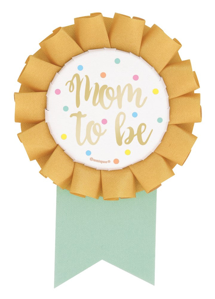 Round badge with gold ruffled edge, white center reading "mom to be" in gold script, pastel confetti, and a mint green.