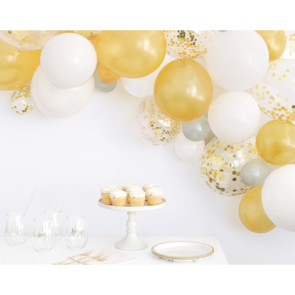 Gold, white, and clear latex balloons with gold confetti arranged in a festive arch over a white table with cupcakes and.
