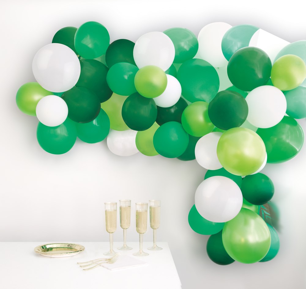 Green, white, and light green balloons arranged in a dense arch cluster above a table with champagne glasses and party.