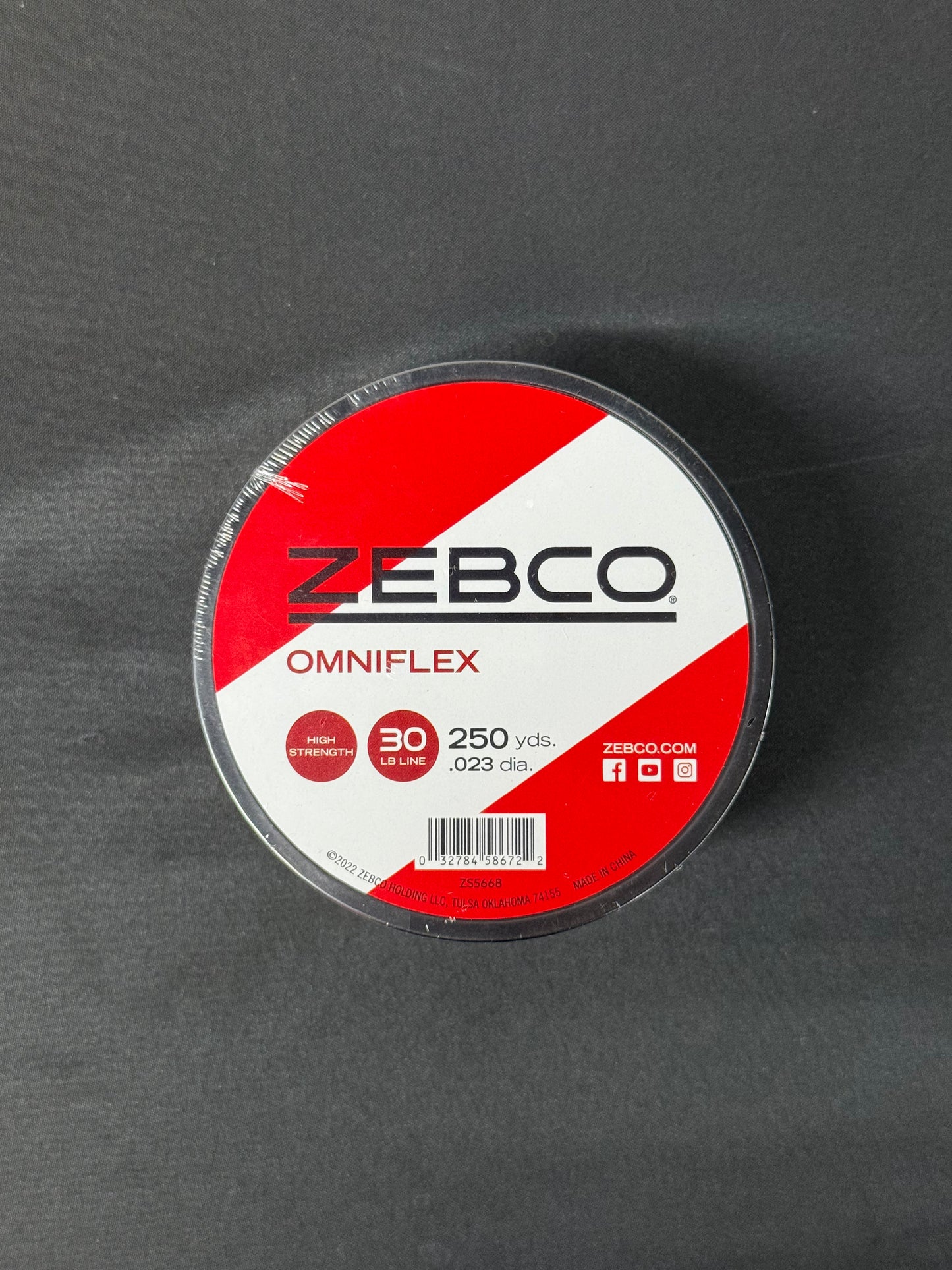 Round spool of Zebco Omniflex 30lb fishing line with red and white label on a dark surface.