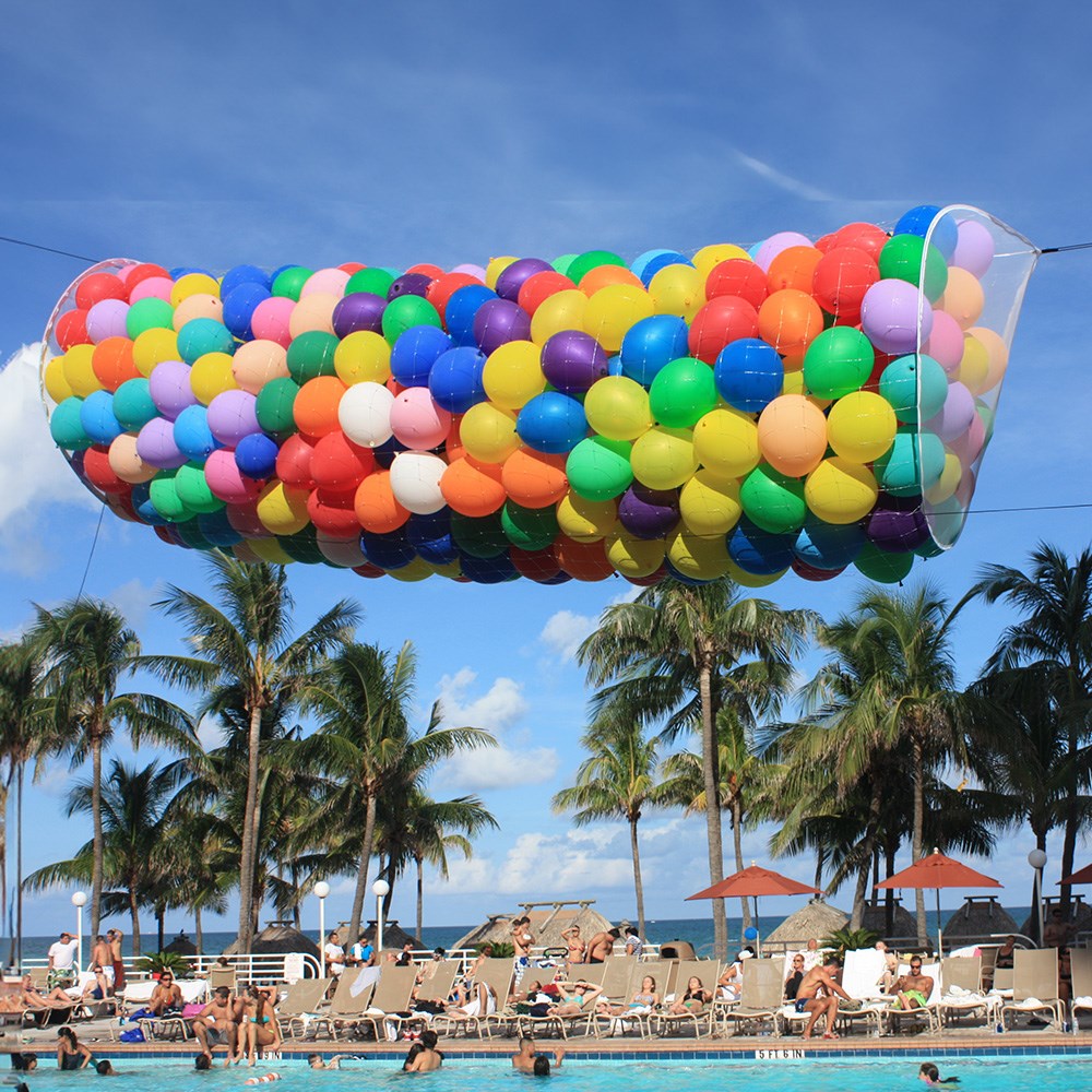 Silver balloon drop net filled with colorful balloons, suspended above outdoor pool area.