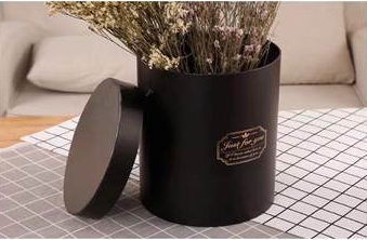 Set of three matte black cylindrical gift boxes with lids, one holding dried flowers and a gold foil label on the front.