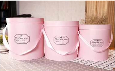 Set of three pink round gift boxes with lids and handles, each featuring a decorative label on the front.