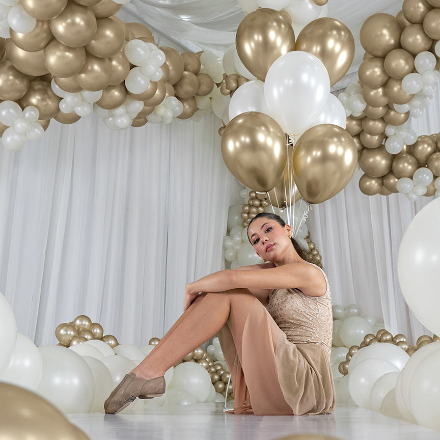 Cluster of shiny gold and white 5-inch latex balloons with a silk finish, held by a woman sitting on the floor surrounded by