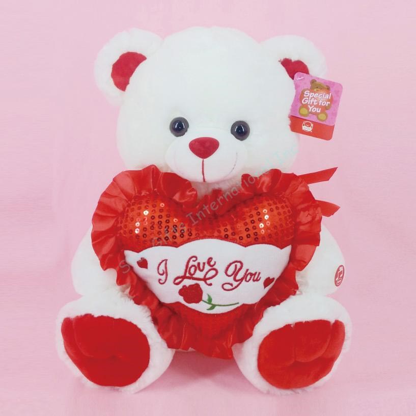 Plush white teddy bear holding red heart pillow with "I Love You" embroidered and red ruffle trim.