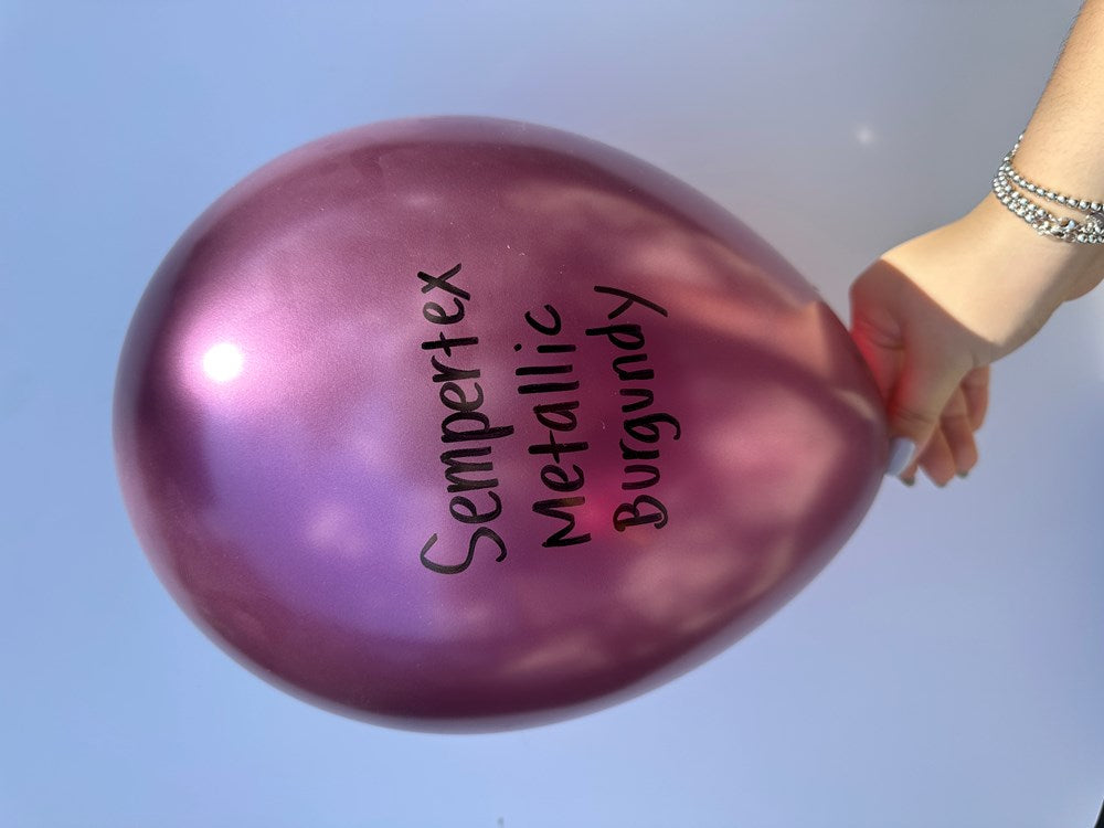 Shiny metallic burgundy latex balloon inflated round held by a hand against a blue sky background.
