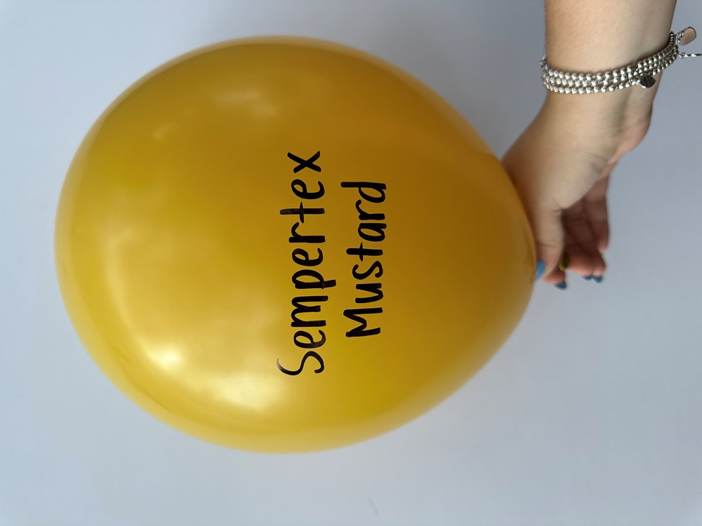 Round mustard yellow latex balloon with black "Sempertex Mustard" text held by a hand.