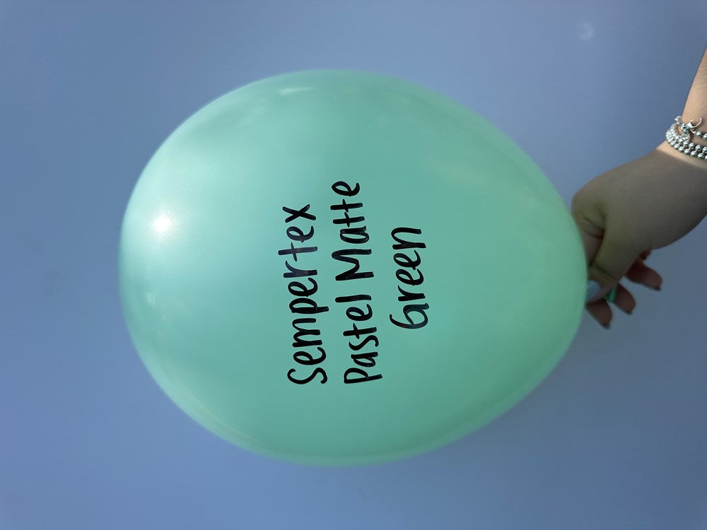 Single pastel matte green latex balloon inflated, round shape, smooth finish, hand holding tied end.