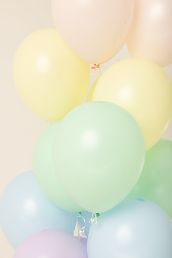 Cluster of pastel-colored latex balloons including yellow, pale pink, light green, and soft blue, floating helium inflated.