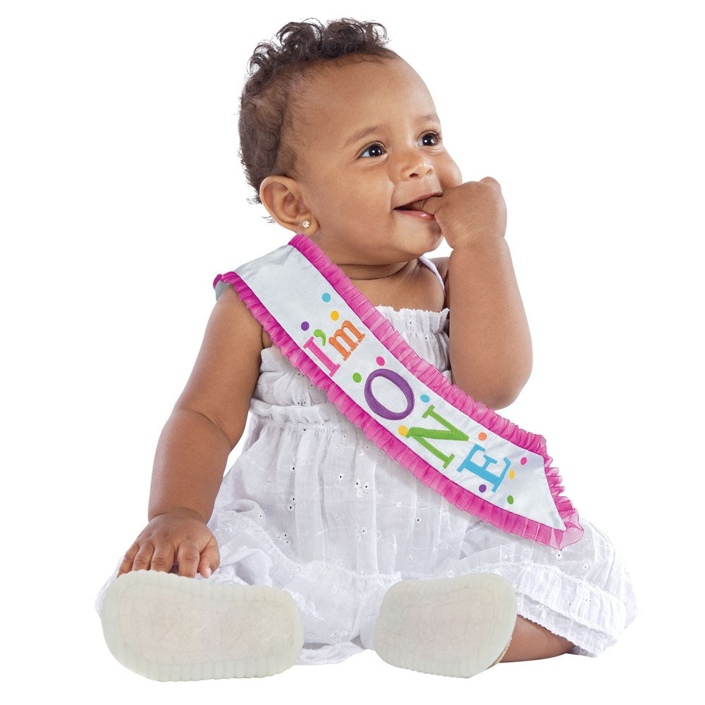 Baby girl wearing a white dress and a pink birthday sash with colorful "One" text.
