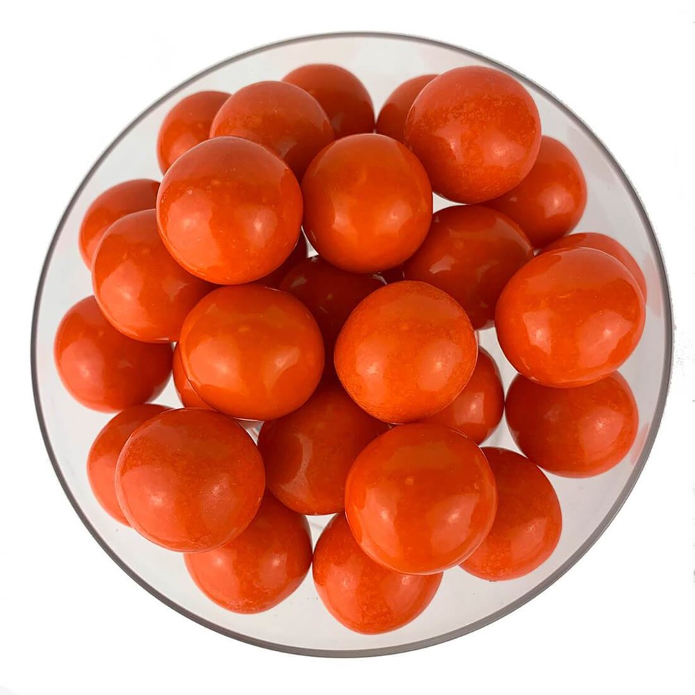 Glossy bright orange round gumballs piled in a clear glass bowl against white background.