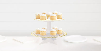 Two-tier white cupcake stand with gold polka dots, holding vanilla cupcakes with white frosting.
