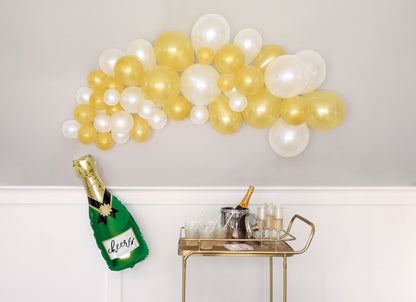 Gold and white latex balloon garland with transparent balloons, above a gold bar cart holding a champagne bottle cooler,.